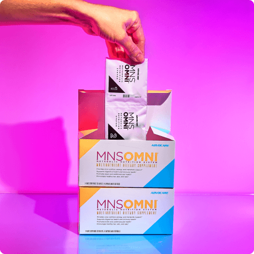 Hand grabbing packets of MNS Omni out of stacked boxes against a pink background.