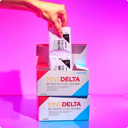 Hand grabbing packets of MNS Delta out of stacked boxes against a pink background.